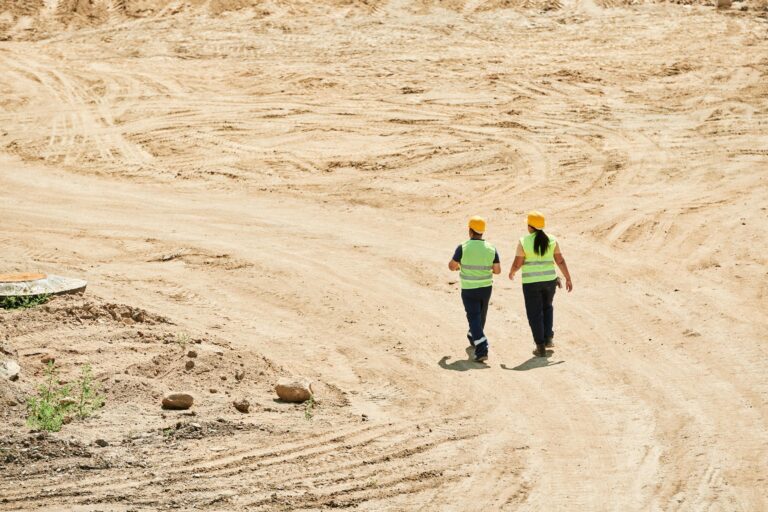 Two construction workers in safety gear walk through a dusty construction site, highlighting teamwork and industry.